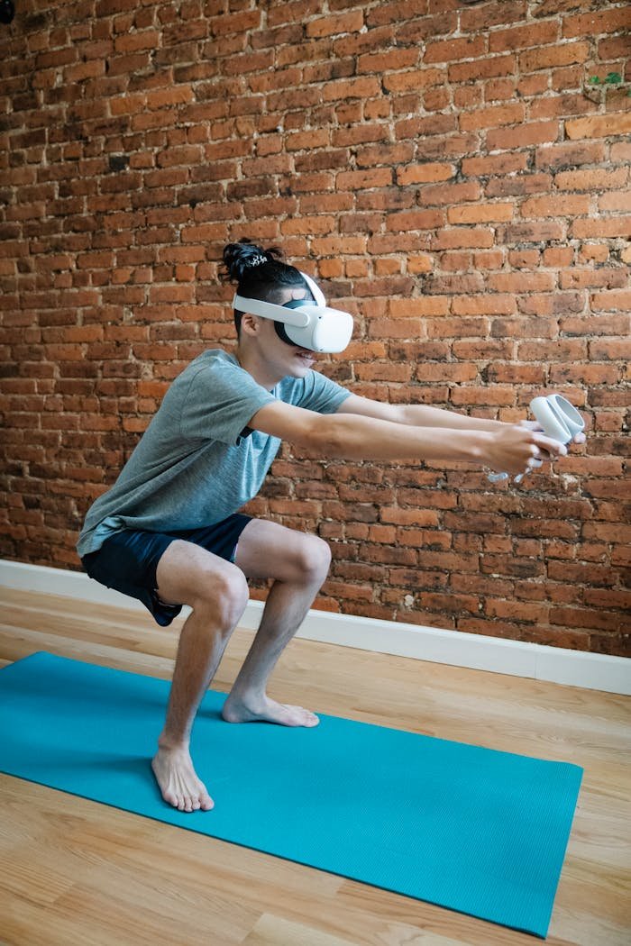 Fit man in virtual reality headset with controllers playing video game while practicing yoga against brick wall at home